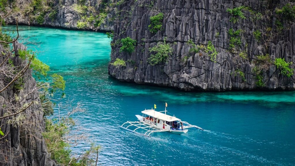3-Day Coron to El Nido Expedition: Is Island Hopping Worth It? 1 Aerial view of a traditional Filipino boat cruising through crystal-clear turquoise waters, surrounded by limestone cliffs and hidden lagoons of Palawan. 3-Day Coron to El Nido Expedition, Island Hopping